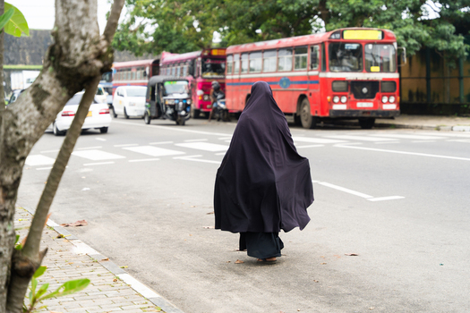 Sri Lanka 15 February 2026 View of a woman in a black niqab walking on a sidewalk near a crosswalk with red buses and a tuk tuk in the background.