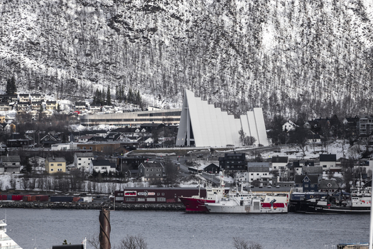 View of the Arctic Cathedral with its iconic triangular architecture against a snowy mountain backdrop and the harbor in Tromso Troms Norway.