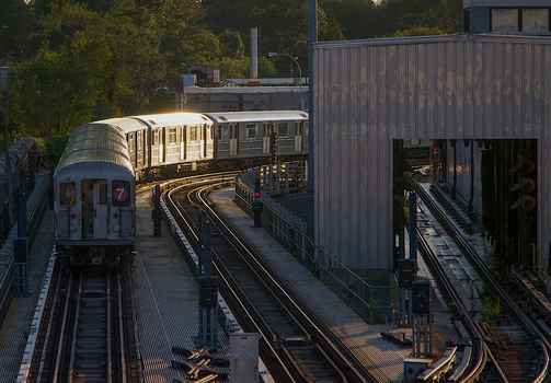 View of a New York City subway train on the 7 line curving along elevated tracks past industrial buildings and green trees during sunset in New York New York United States. View of a New York City subway train on the 7 line curving along elevated tracks past industrial buildings and green trees during sunset in New York New York United States.
