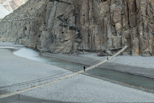 View of Hussaini Suspension Bridge with people walking across the wooden planks over the river set against rugged mountain cliffs in Hussaini Gilgit Pakistan. View of Hussaini Suspension Bridge with people walking across the wooden planks over the river set against rugged mountain cliffs in Hussaini Gilgit Pakistan.