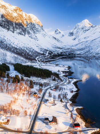 Aerial view of the serpentine road snaking along the fjord framed by snow laden mountains under a crisp blue sky Indregård Troms Norway. Aerial view of the serpentine road snaking along the fjord framed by snow laden mountains under a crisp blue sky Indregård Troms Norway.