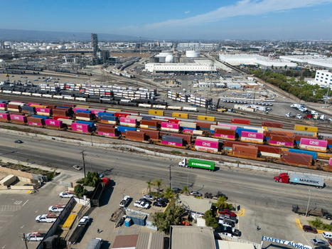Aerial view of the industrial rail yard in Carson with colorful shipping containers on freight trains adjacent to a highway with trucks and a refinery complex Carson California United States.