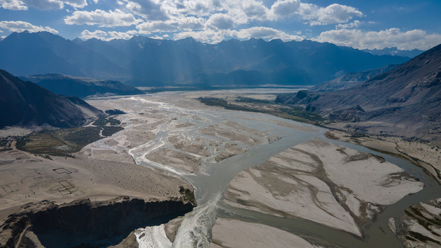 Aerial view of the Shigar River flowing through a vast sandy valley surrounded by rugged snow capped mountains under a bright cloudy sky in Skardu Gilgit Pakistan.