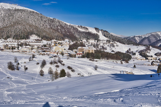 View of a snowy valley with ski tracks leading towards buildings nestled beneath towering mountains under a clear blue sky Donovaly Banska Bystrica Region Slovakia. View of a snowy valley with ski tracks leading towards buildings nestled beneath towering mountains under a clear blue sky Donovaly Banska Bystrica Region Slovakia.