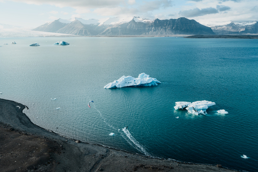 Aerial view of Jokulsarlon glacier lagoon with floating icebergs and a kitesurfer gliding across turquoise water near the dark volcanic shore in Sveitarfelagid Hornafjordur Iceland. Aerial view of Jokulsarlon glacier lagoon with floating icebergs and a kitesurfer gliding across turquoise water near the dark volcanic shore in Sveitarfelagid Hornafjordur Iceland.