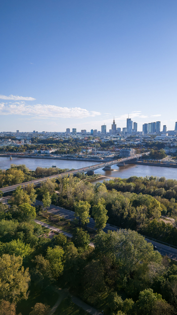 Aerial view of the Vistula River flowing serenely beneath a bridge amidst lush green trees and the distant Warsaw skyline Warsaw Poland. Aerial view of the Vistula River flowing serenely beneath a bridge amidst lush green trees and the distant Warsaw skyline Warsaw Poland.
