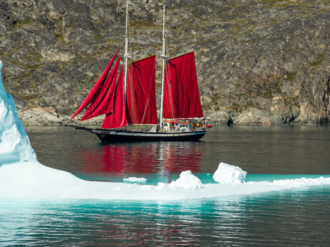 Aerial view of a sailboat with striking red sails navigating through calm icy waters past a large iceberg with a rugged rocky coastline in the background in Greenland. Aerial view of a sailboat with striking red sails navigating through calm icy waters past a large iceberg with a rugged rocky coastline in the background in Greenland.