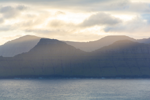 View of mountains rising above the ocean under a cloudy sky in the Faroe Islands. View of mountains rising above the ocean under a cloudy sky in the Faroe Islands.