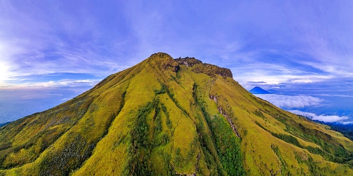 Aerial view of the lush green slopes and rugged peak of Mount Sumbing under a clear blue sky with a sea of clouds in the distance Temanggung Central Java Indonesia. Aerial view of the lush green slopes and rugged peak of Mount Sumbing under a clear blue sky with a sea of clouds in the distance Temanggung Central Java Indonesia.