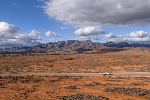 Aerial view of rugged mountain peaks and vast outback desert landscape with a white vehicle driving on a road under a cloudy sky in Flinders Ranges South Australia Australia. Aerial view of rugged mountain peaks and vast outback desert landscape with a white vehicle driving on a road under a cloudy sky in Flinders Ranges South Australia Australia.