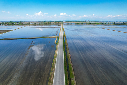 Aerial view of flooded rice fields reflecting the blue sky with a straight road leading towards industrial cooling towers in the distance Trino Piemonte Italy.