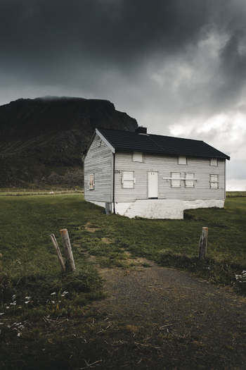 View of Haukland Beach featuring a white wooden house with boarded windows set against a dramatic dark mountain under a cloudy sky in Leknes Nordland Norway. View of Haukland Beach featuring a white wooden house with boarded windows set against a dramatic dark mountain under a cloudy sky in Leknes Nordland Norway.