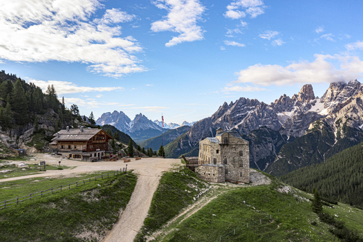 View of Prato Piazza featuring historic stone and wooden mountain huts against the jagged peaks of the Dolomites under a blue sky in Dobbiaco Trentino Alto Adige Italy. View of Prato Piazza featuring historic stone and wooden mountain huts against the jagged peaks of the Dolomites under a blue sky in Dobbiaco Trentino Alto Adige Italy.