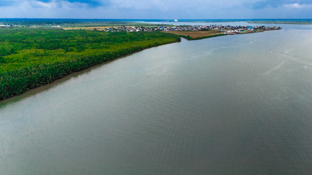 Aerial view of a river flowing past a small settlement surrounded by dense green vegetation under a cloudy sky in Minima Rivers Nigeria.
