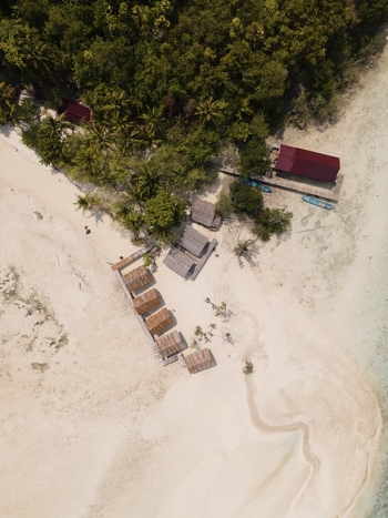 Aerial view of Ransiwor homestay featuring thatched roof huts on a pristine white sand beach surrounded by lush tropical vegetation and turquoise waters in Raja Ampat Southwest Papua Indonesia. Aerial view of Ransiwor homestay featuring thatched roof huts on a pristine white sand beach surrounded by lush tropical vegetation and turquoise waters in Raja Ampat Southwest Papua Indonesia.