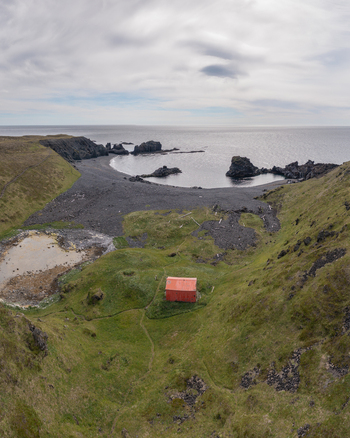 Aerial view of Djupalonssandur featuring a small red building nestled in grassy hills overlooking a dramatic black sand beach and rocky coastline in Iceland. Aerial view of Djupalonssandur featuring a small red building nestled in grassy hills overlooking a dramatic black sand beach and rocky coastline in Iceland.