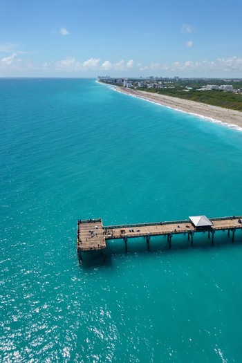 Aerial view of Juno Beach Pier extending into turquoise ocean water along the sandy coastline under a clear blue sky Juno Beach Florida United States. Aerial view of Juno Beach Pier extending into turquoise ocean water along the sandy coastline under a clear blue sky Juno Beach Florida United States.