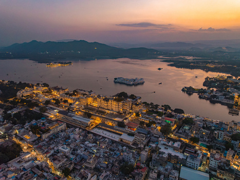Aerial view of the illuminated City Palace and Lake Palace on Lake Pichola at sunset with the city skyline and distant mountains in Udaipur Rajasthan India.