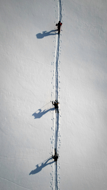 Aerial view of three people trekking a snowy path their long shadows stretching behind them across the pristine white expanse Anniviers Valais Switzerland.