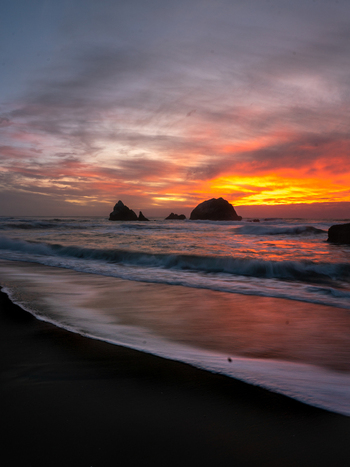 View of a vibrant sunset over the ocean with sea stacks rising from the water and waves washing onto the sandy shore in United States of America. View of a vibrant sunset over the ocean with sea stacks rising from the water and waves washing onto the sandy shore in United States of America.