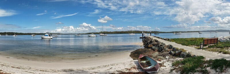 Aerial view of boats bobbing gently on the water a pier stretches into the calm bay and a rustic boat sits on the sandy shore Forster Australia.