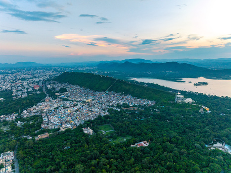 Aerial view of the city of Udaipur nestled between lush green hills and the serene waters of Lake Pichola at sunset featuring dense urban architecture in Udaipur Rajasthan India.