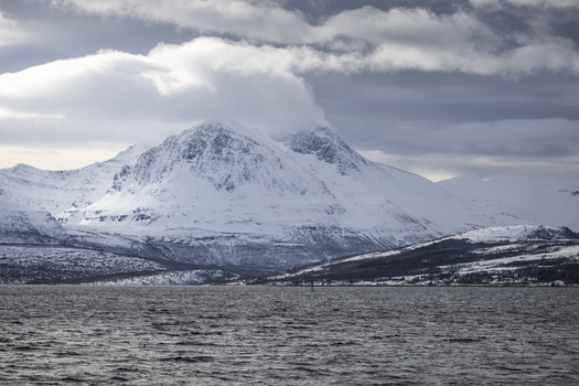 View of snow capped mountains and a calm fjord under a dramatic cloudy sky in Tromso Troms Norway.