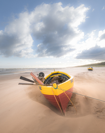 View of a traditional yellow and red wooden fishing boat resting on a sandy beach under a cloudy sky with blowing sand in Debki Pomeranian Voivodeship Poland. View of a traditional yellow and red wooden fishing boat resting on a sandy beach under a cloudy sky with blowing sand in Debki Pomeranian Voivodeship Poland.