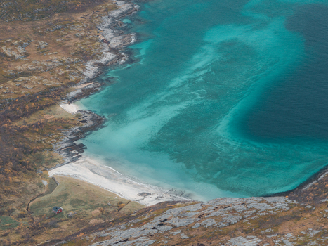 Aerial view of the turquoise waters and sandy beach with rocky terrain and a small red house in Senja Troms Norway.