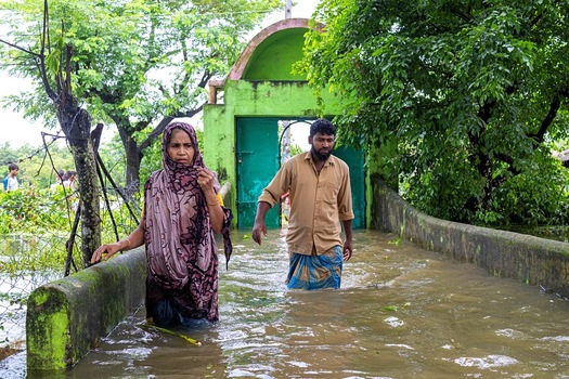 Banshkhali Upazila Chittagong Division Bangladesh 10 July 2026 View of a man and a woman wading through floodwaters in front of a green gate in a rural area. Banshkhali Upazila Chittagong Division Bangladesh 10 July 2026 View of a man and a woman wading through floodwaters in front of a green gate in a rural area.