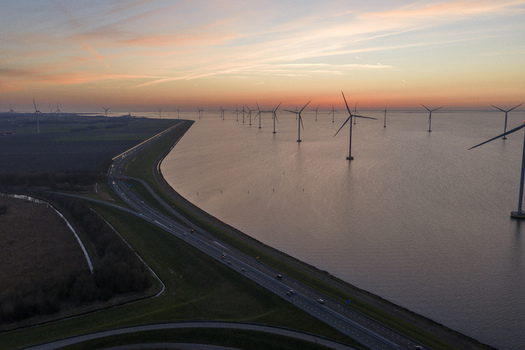 Aerial view of the IJsselmeer wind farm with turbines standing in the water next to a highway on a dike during a vibrant sunset in Netherlands.