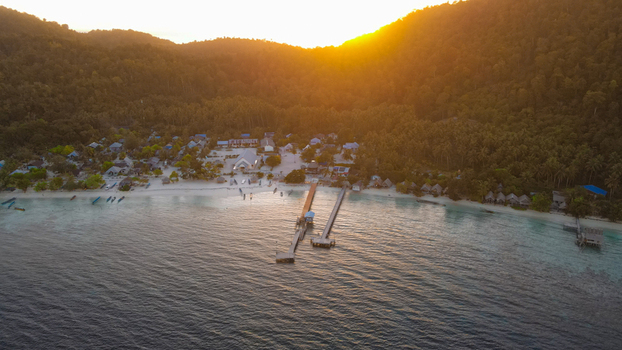 Aerial view of Raja Ampat coastal village with wooden piers extending into the clear water surrounded by lush tropical hills at sunset in Raja Ampat Southwest Papua Indonesia.