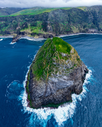 Aerial view of the stark contrast of the rugged rock of the islet against the deep blue ocean with the green cliffs in the background Flores A&ccedil;ores Portugal.