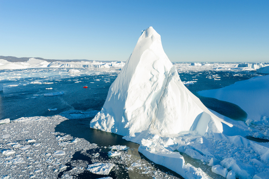 Aerial view of Ilulissat glacier with a massive white iceberg floating in the deep blue sea surrounded by smaller ice fragments and a small red boat Greenland. Aerial view of Ilulissat glacier with a massive white iceberg floating in the deep blue sea surrounded by smaller ice fragments and a small red boat Greenland.