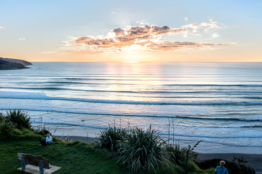 Raglan New Zealand 03 September 2021 Aerial view of the coastline at sunset with gentle waves rolling onto the beach lush green vegetation on the cliffside and a person on a bench.