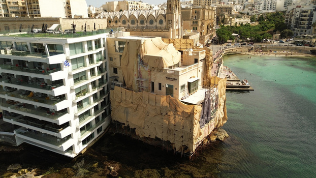 Aerial view of a historic building under renovation covered in protective sheets next to a modern hotel overlooking the turquoise waters of a bay in Malta. Aerial view of a historic building under renovation covered in protective sheets next to a modern hotel overlooking the turquoise waters of a bay in Malta.