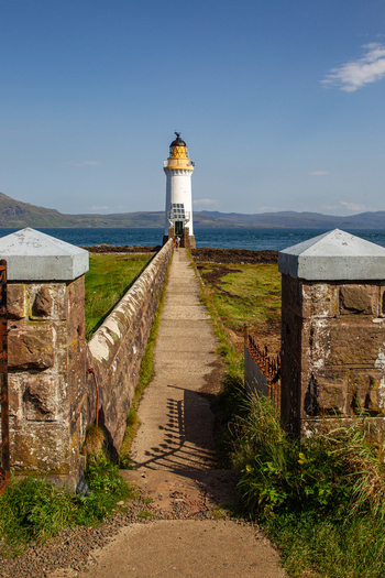 View of Tobermory Lighthouse at the end of a stone walkway framed by pillars with the sea and hills in the background in Isle of Mull Scotland United Kingdom. View of Tobermory Lighthouse at the end of a stone walkway framed by pillars with the sea and hills in the background in Isle of Mull Scotland United Kingdom.