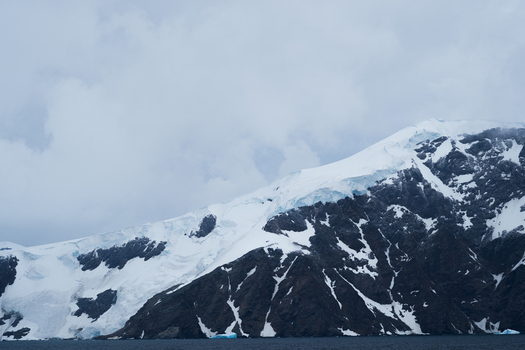 View of the snow covered mountains and glaciers with dark rocky slopes and a cloudy sky above the sea in the South Orkney Islands. View of the snow covered mountains and glaciers with dark rocky slopes and a cloudy sky above the sea in the South Orkney Islands.