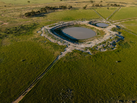 Aerial view of a circular water dam surrounded by green pasture and dirt tracks in the flat landscape of Hay New South Wales Australia.