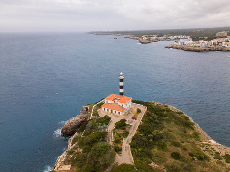 Aerial view of Far de Portocolom lighthouse on a rocky cliff overlooking the blue sea and coastal town Portocolom Balearic Islands Spain. Aerial view of Far de Portocolom lighthouse on a rocky cliff overlooking the blue sea and coastal town Portocolom Balearic Islands Spain.