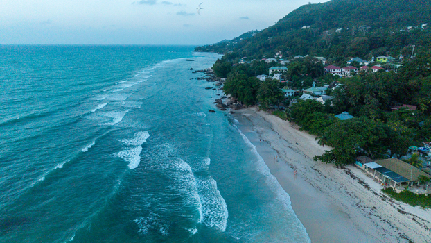 Aerial view of Beau Vallon Beach with turquoise waves crashing onto the sandy shore surrounded by lush green hills and coastal houses in Beau Vallon Seychelles.