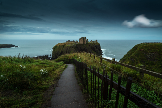 View of the historic Dunnottar Castle ruins on a rocky headland overlooking the North Sea with a coastal path and wooden fence under a moody sky in Scotland.