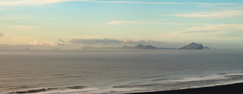 Aerial view of the Westman Islands rising from the sea under a soft sky with calm waves washing onto the black sand beach in Hvolsvollur Rangarthing eystra Iceland.