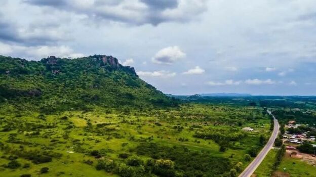 Aerial view of a lush green hill rising above a vast landscape with a winding road and scattered vegetation under a cloudy sky in Ghana. Aerial view of a lush green hill rising above a vast landscape with a winding road and scattered vegetation under a cloudy sky in Ghana.