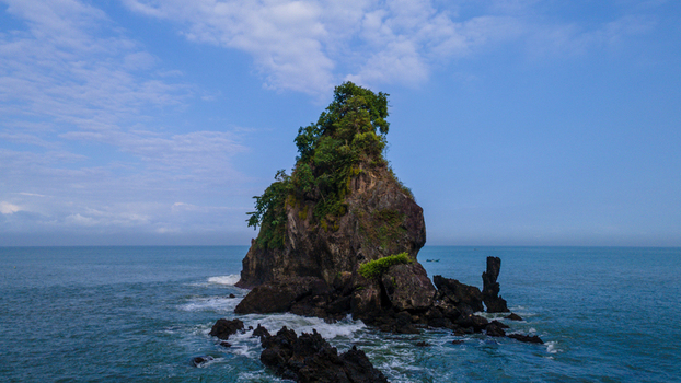 Aerial view of the iconic Karang Agung Beach rock formation covered in lush vegetation surrounded by the blue ocean and dark rocks in Ayah Central Java Indonesia. Aerial view of the iconic Karang Agung Beach rock formation covered in lush vegetation surrounded by the blue ocean and dark rocks in Ayah Central Java Indonesia.