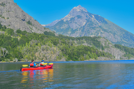 View of two people in a red kayak on the calm waters of Nahuel Huapi Lake with lush green forests and a mountain peak San Carlos de Bariloche Rio Negro Argentina. View of two people in a red kayak on the calm waters of Nahuel Huapi Lake with lush green forests and a mountain peak San Carlos de Bariloche Rio Negro Argentina.