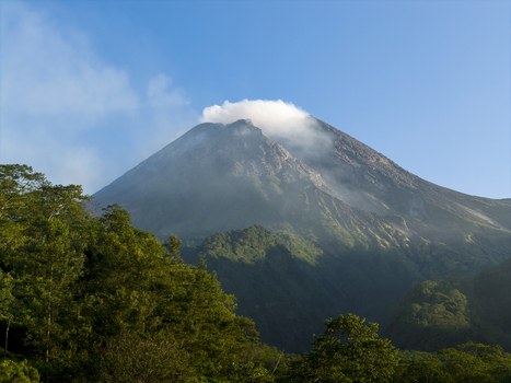 Aerial view of Mount Merapi volcano with smoke rising from the crater above lush green forests and hills in Kecamatan Pakem Daerah Istimewa Yogya Indonesia. Aerial view of Mount Merapi volcano with smoke rising from the crater above lush green forests and hills in Kecamatan Pakem Daerah Istimewa Yogya Indonesia.