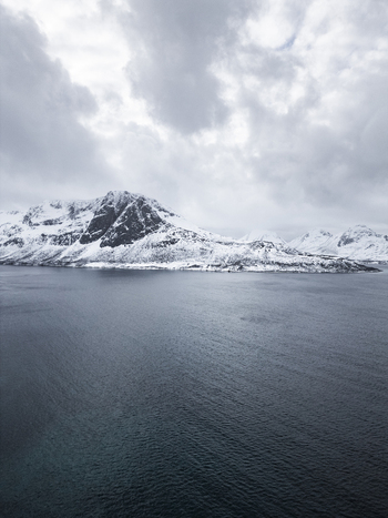 Aerial view of Grotfjord with snow covered mountains rising from the dark blue sea under a cloudy sky in Tromvik Troms Norway. Aerial view of Grotfjord with snow covered mountains rising from the dark blue sea under a cloudy sky in Tromvik Troms Norway.