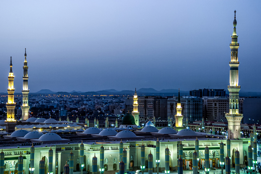 View of Al Masjid e Nabawi at dusk with illuminated minarets and domes set against the city skyline of Madinah Al Madinah Province Saudi Arabia.