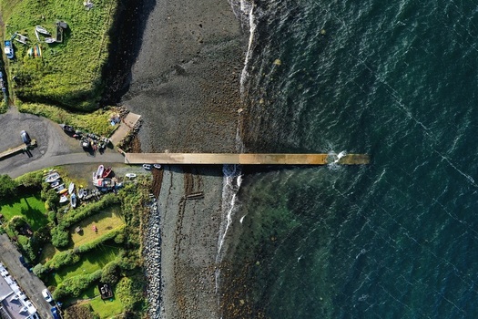 Aerial view of a concrete pier extending into the dark blue sea from a rocky shoreline with small boats and green grassy banks in Stein Scotland United Kingdom. Aerial view of a concrete pier extending into the dark blue sea from a rocky shoreline with small boats and green grassy banks in Stein Scotland United Kingdom.